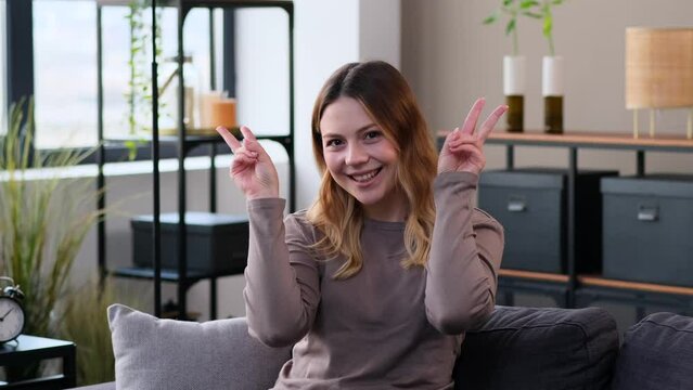 Smiling and friendly Caucasian woman showing victory or peace hand sign sitting on sofa at home living room. Symbol or gesture of a winner.