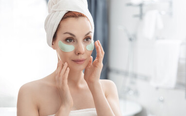 Woman wrapped in towel is examining her face in mirror, focusing on her skin, lips, eyebrows. She smiles at her reflection. Girl with towel on head takes care of her facial skin