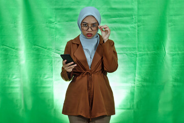unhappy young Asian woman wearing hijab, glasses and blazer holding smartphone and eyewear looking...