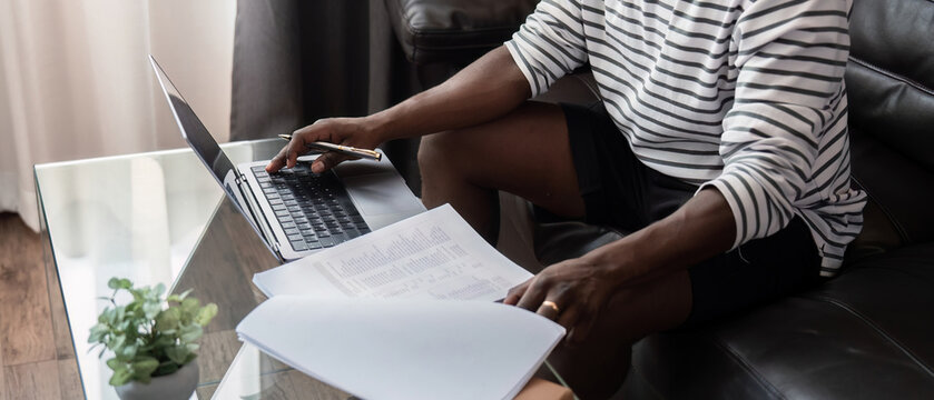 African American man working with laptop computer remote while sitting at sofa in living room. Black guy do freelance work at home office