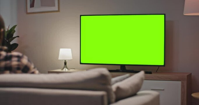 Man sitting at sofa and enjoying favourite TV show. Back view of male sitting in front of TV. African American man relaxing sitting at couch at home. Male looking at green chroma key screen of TV.