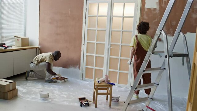 Rear full footage of diverse wife and husband painting walls brown using roller and brush during renovation process in new apartment