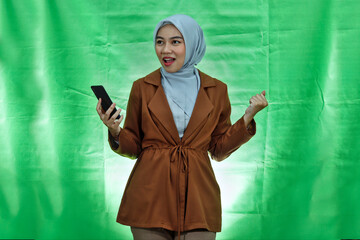excited young Asian woman wearing hijab and blazer holding smartphone and celebrating success over...