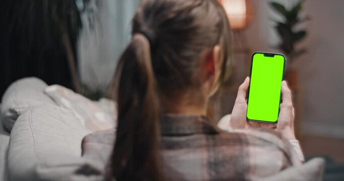 Woman sitting at couch watching green screen. Female watching chroma key screen at blurred background. Woman holding smartphone and watching series. Lady in living room starring at gadget.