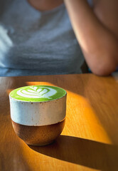 Cup of matcha latte green tea on wooden table with woman in background.