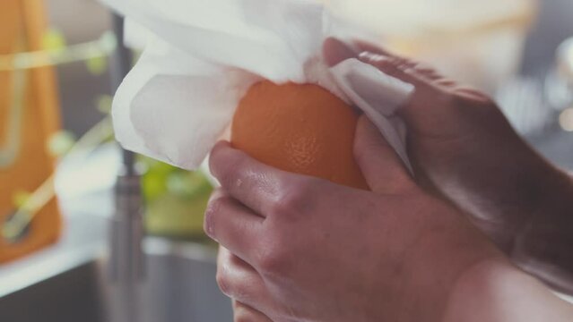Close up of woman's hands cleaning, and wiping dry a whole fresh lemon with paper towel. Defocused brightly lit kitchen in the background. Studio shot. . High quality 4k footage