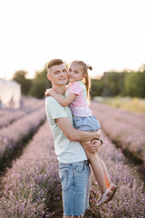 Father hugs daughter in flowers lavender enjoying scent, on summer day at sunset. Dad holds hands child girl on lavender field background. Daddy and baby embrace in meadow. Happy family outdoors.