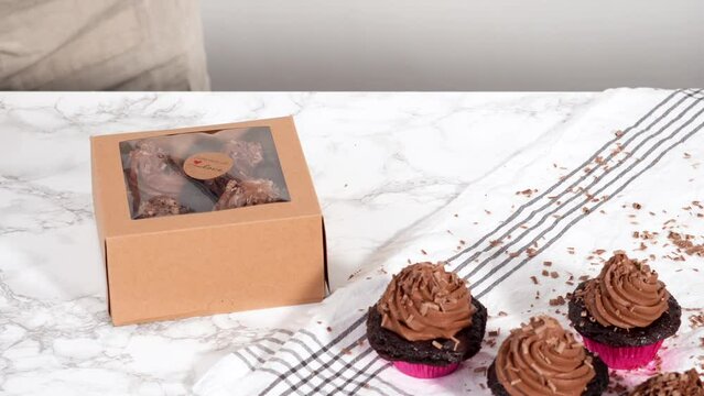 Packaging chocolate cupcakes with chocolate ganache frosting into a paper cupcake box.