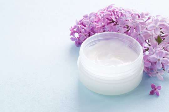 Fresh purple pink lilac flowers and plastic white cream jar on light blue table background. Care about face, hands, legs and body skin. Closeup. Front view.