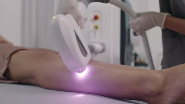 Close up of hands of beautician performing laser epilation treatment on legs of female client in spa salon