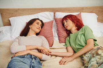 happy young lesbian couple looking at each other while lying together on bed, holding hands