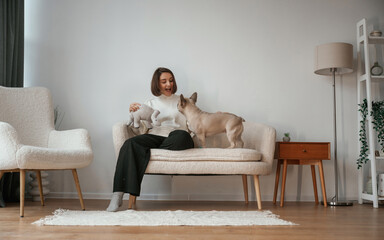 Front view, sitting on the little sofa. Young woman is with her pug dog at home