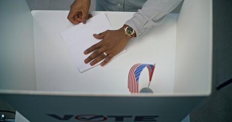 Close up of African American man filling out paper bulletin in voting booth with American flag....