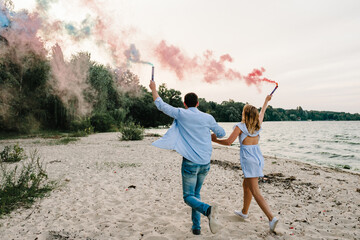 Man and woman having fun running on sand and holding colorful blue and red or pink smoke bombs on...