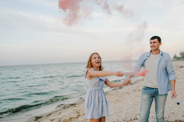 Man and woman having fun standing on sand and holding colorful red, pink color smoke bombs and look...