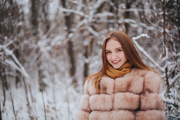 Attractive young brunette woman wearing fur coat with scarf standing alone in front of snowy forest