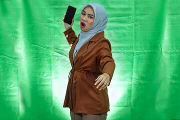 angry young Asian woman wearing hijab and blazer with displeased expression and wanna throw mobile phone over green background
