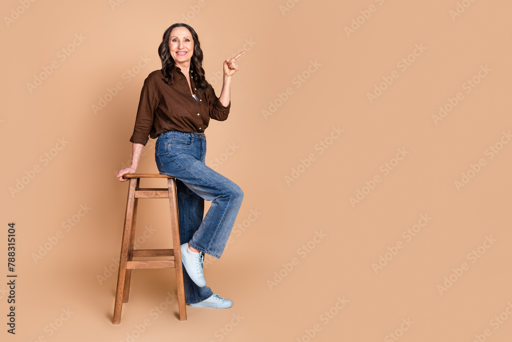 Sticker Full size photo of attractive senior woman sit chair point empty space dressed stylish brown clothes isolated on beige color background