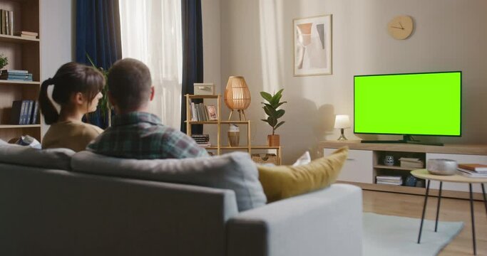 Young couple spending time together sitting at couch. Man and woman watching green chroma key TV. Married couple sitting at sofa and talking. Male and female relaxing at home at weekend.