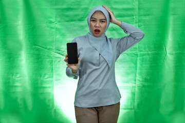 beautiful asian woman wearing hijab and blouse holding smartphone touching her head and expressing concern on green background
