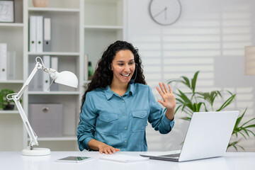Friendly businesswoman in a smart blue shirt, waving and smiling during a virtual meeting in a...