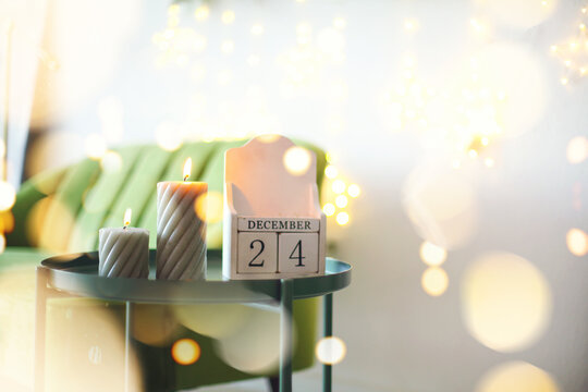December 24 on wooden calendar standing on small table with burning candles in living room, selective focus. Christmas and New Year concept