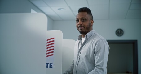 African American man fills out paper ballot in voting booth, than smiles and looks at camera. US...