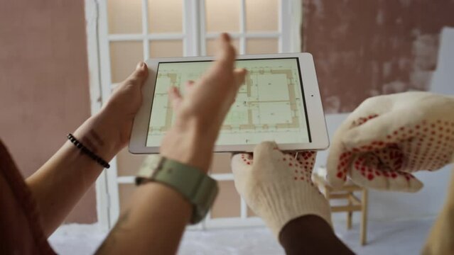 Over shoulder footage of hands in gloves of anonymous married couple pointing at home remodeling plan on digital tablet while discussing new apartment renovation