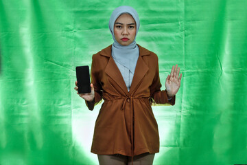 surprised young Asian woman wearing hijab and blazer open mouth showing blank screen smartphone and doing stop gesture with palms over green background.
