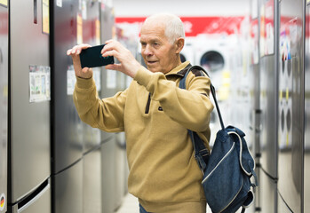 Elderly man taking photo of refrigerator in showroom of electrical appliance store