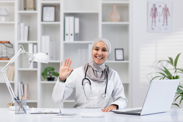 A cheerful female doctor in hijab waves to the camera while sitting at a desk in a bright, well-organized office. Technology and healthcare professionalism combined.