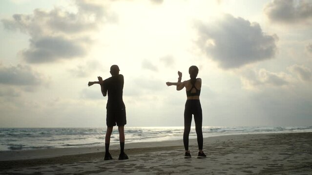 Back side couple young two friends stretching arm before running at seaside. Young couple wearing sportswear running on sand sea ocean beach outdoor jog on seaside. Training athlete work out outdoor.
