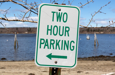Old two hour parking sign with a left arrow at an angle in front of a beach and ocean in the springtime in New England. - 788347126