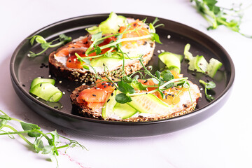 Salmon sandwiches with fullgrain bread, pea micro greens, cream cheese and cucumbers on white marble background close up.
