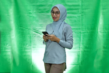 cheerful young Asian woman wearing hijab, glasses and blouse using a mobile phone and gesturing thumbs up isolated on white background

