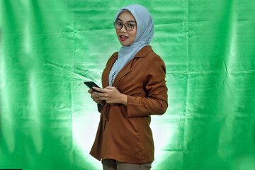 smiling young Asian woman wearing hijab, glasses and blazer using mobile phone isolated on green background
