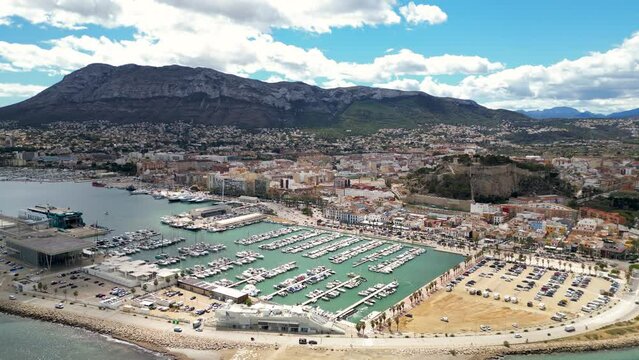 Panoramic aerial view of Denia city. View of port area. Luxury yachts. Balearic Ferry docked in the port. Castle of Denia in the middle of the city. Drone forwards.  