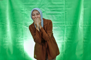 laughing young Asian woman wearing hijab and blazer covering mouth with hand on green background

