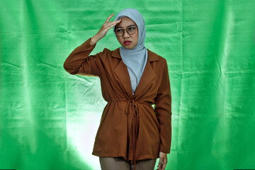 young Asian woman wearing hijab, glasses and blazer looking dizzy and having headache on green background
