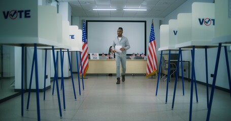 African American male voter walks for registration table at polling station, takes paper ballot. Voting booths with US flag logo and sign calling to vote. Election Day in the United States of America. - obrazy, fototapety, plakaty