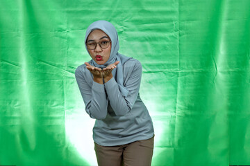 charming young Asian woman wearing hijab, glasses and blouse hold hand palms send air kiss to boyfriend over green background
