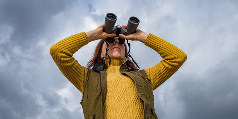 female ornithologist birdman or explorer watches birds with binoculars against a background of a stormy sky - 788369715