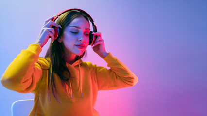 Portrait of a calm Caucasian woman wearing a yellow hoodie sitting and wearing wireless headphones, and posing isolated on a neon light background. Enjoying new albums of favorite music and artists. - 788370111