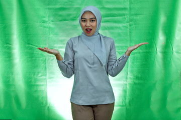 smiling young Asian woman wearing hijab and blouse showing copy space with both palms isolated on green background
