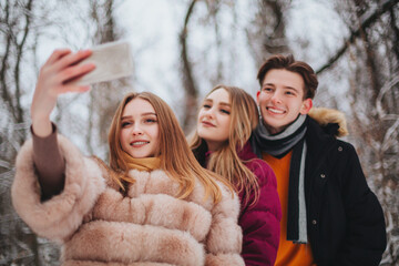 Obraz premium Three young happy friends, two girls and boy making selfie on mobile phone against winter landscape