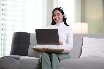Young asian woman using computer while seated on sofa at home. Happy girl wear headphones relaxing...