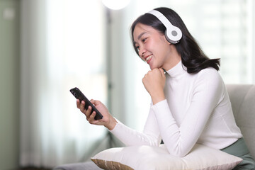 Young asian woman using smartphone, wearing headphone sitting on sofa at home. Happy girl relaxing comfortable in the living room, spending lazy weekend and enjoy listening to music, watching movie.