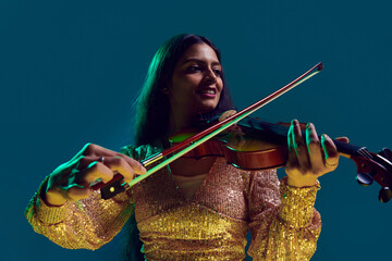 Smiling Indian female violinist in shimmering crop top with serene expression in neon light against...