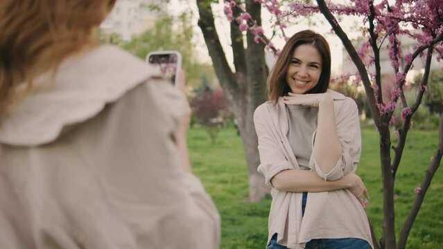 two incredibly beautiful young women filming each other on a smartphone in the park against the background of pink blooming spring trees. Girls laugh, rejoice and make faces at the camera