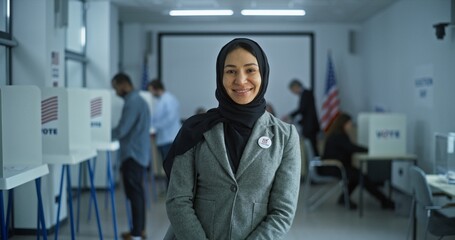 Portrait of Muslim woman, United States of America elections voter. Woman stands in a modern polling station, poses, smiles and looks at camera. Background with voting booths. Concept of civic duty.